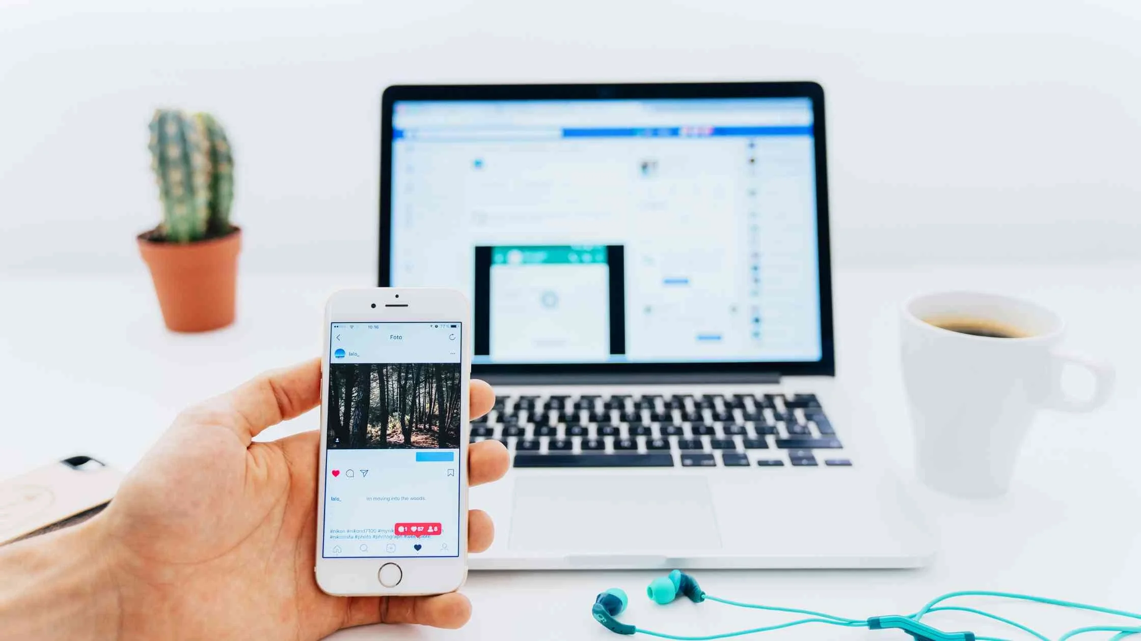 Person holding a smartphone displaying an Instagram feed, with a laptop, coffee, and cactus on a white desk.